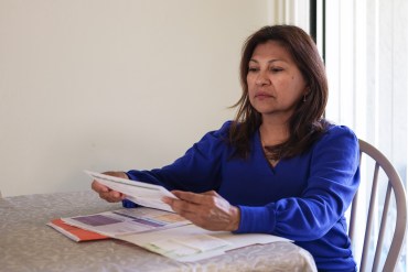 A Hispanic woman sits at a table, reading medical bills from a red file folder in front of her.