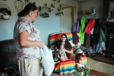 A doula stands next to a young mother sitting in a chair draped with colorful fabric. She holds a young baby in her arms.