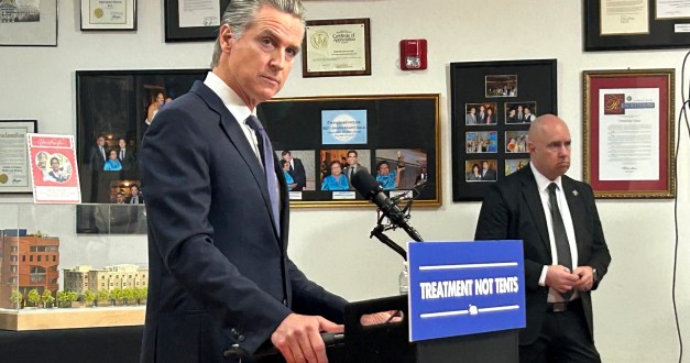Gavin Newsom speaks at a news conference behind a podium with a sign on it that reads, "Treatment not tents."