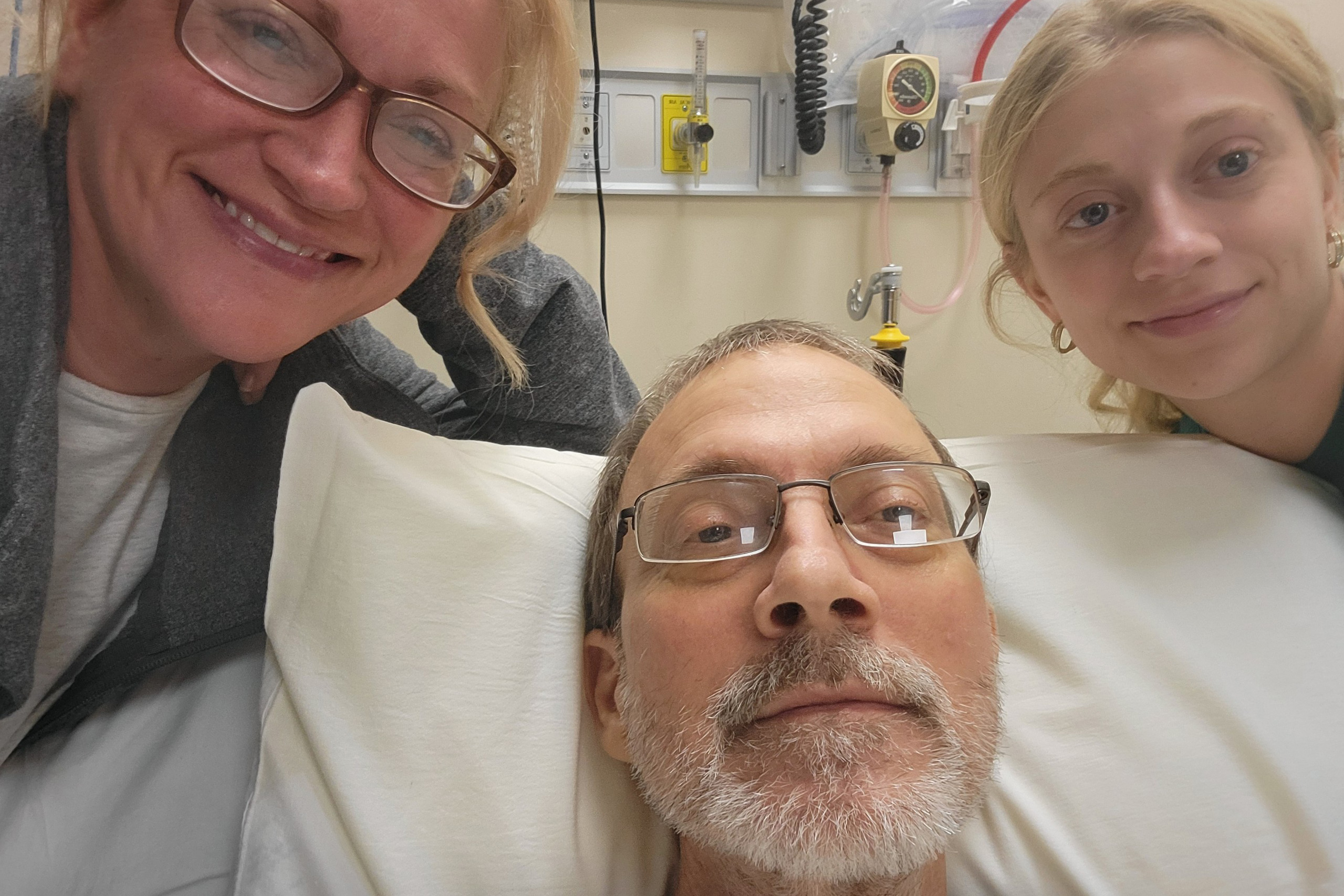 An older man lies in a hospital bed. His wife and daughter are on either side of him, smiling.